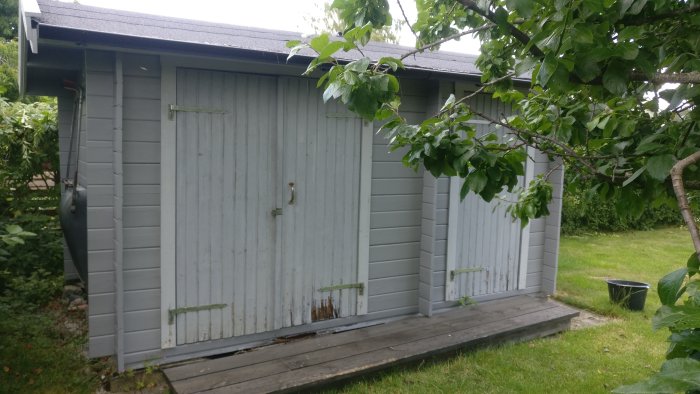 Grått trädgårdsförråd med skadad bräda längst ned nära lecablocket, synliga tecken på förfall.
