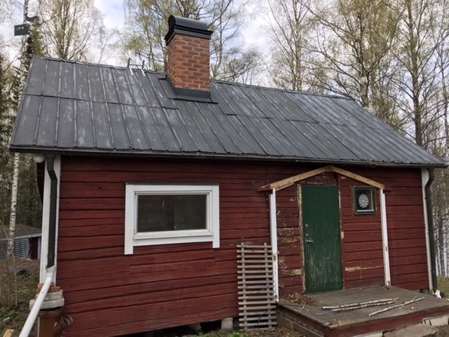 Exteriör av timrat torp med röd liggande timmerpanel, gröna dörrar och plåttak.