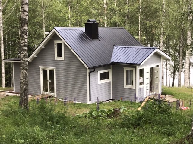 Moderniserat grått hus med liggande panel och svart plåttak omgivet av björkar, efter renovering av gammalt timrat torp.