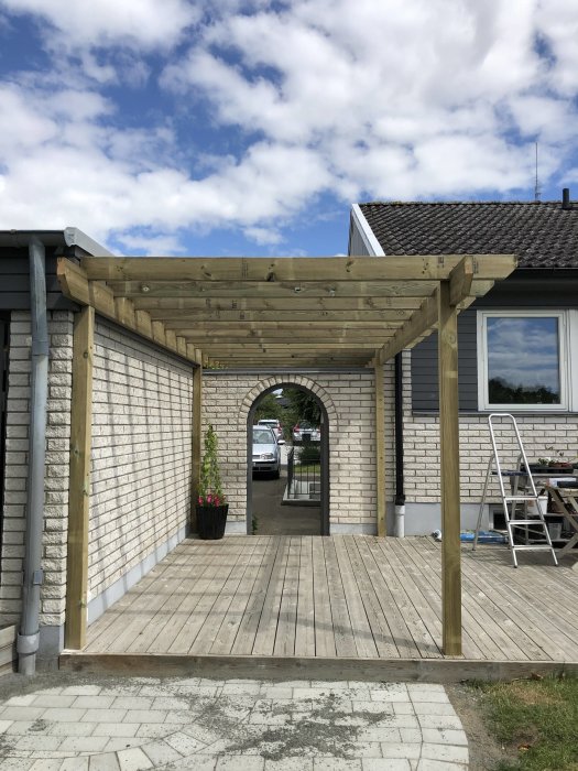 Nybyggd träpergola på en trädäck framför ett hus, under en klarblå himmel med moln.