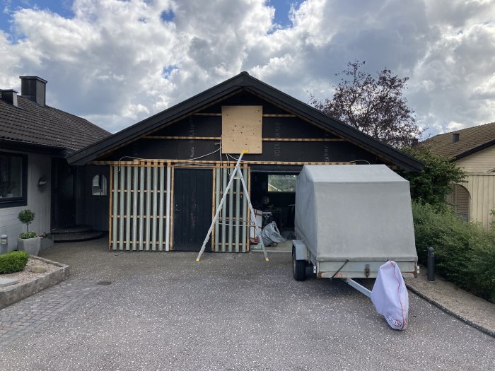 Garage med delvis borttagen fasad och synlig isolering, en stege lutar mot bygget, släpvagn framför.