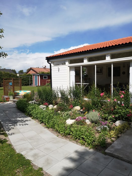 Välansat blomsterbed med rosor och stenplattor framför ett vitt hus, möjlig plats för framtida smidesstaket.