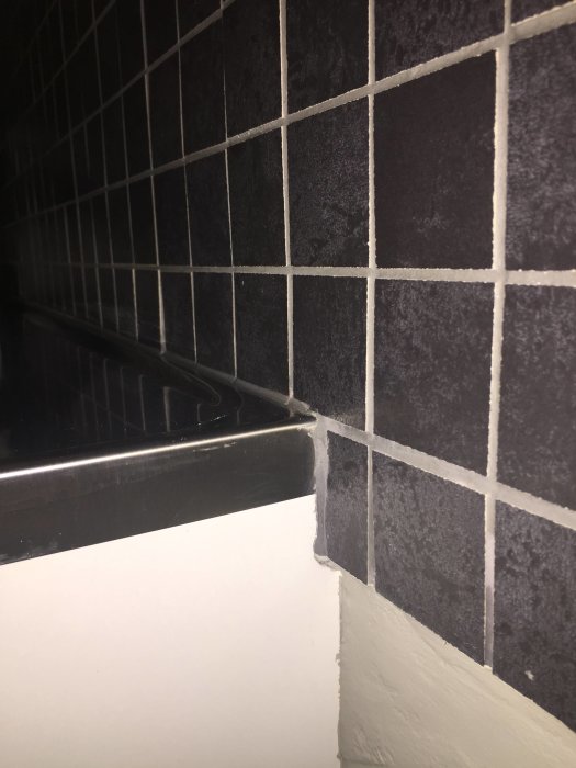 Mörka kakelplattor ovanför rostfri diskbänk utan uppvik, vit fog, möte med vit vägg.