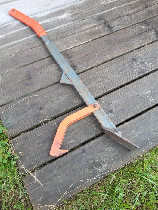 Ett rostigt brytjärn med orange handtag och aggressivt vinklad krok ligger på en trätrall.