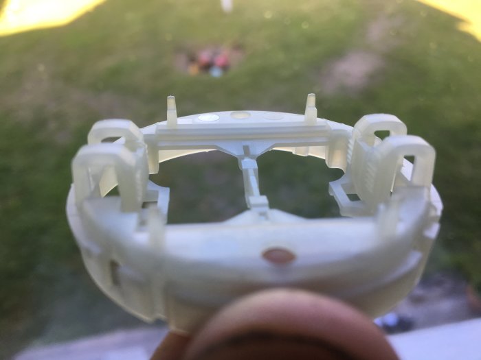 Närbild av vit plastdetalj med rektangulära hål, otydlig bakgrund med gräsmatta och lekande barn.