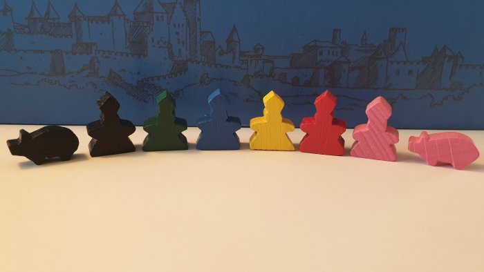 Flerfärgade Carcassonne-brädspelspjäser i rad framför tecknad stadssilhuett, inklusive handgjorda biskop och gris.