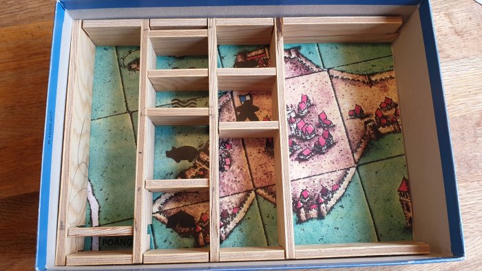 Träinsats med fack i en spelkartong för brädspelstillbehör, på en bakgrund av spelbrickor.