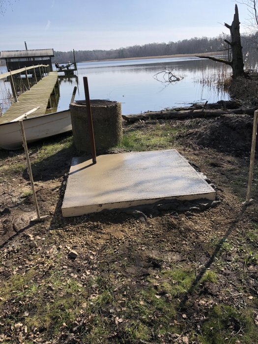 Nybyggd grillplats med betongplatta vid sjö med brygga och båt i bakgrunden.