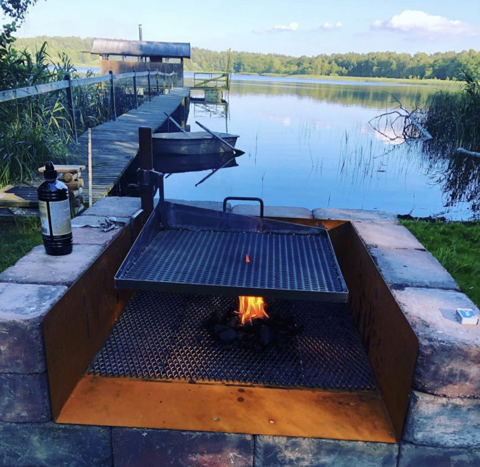 Utomhusgrill vid sjökant med öppen eld på galler och rostfritt grillgaller ovan, i naturlig omgivning.