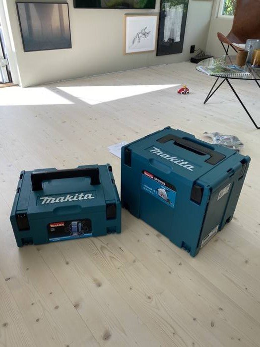 Två Makita verktygslådor står på ett ljust trägolv inne i ett rum med modern inredning i bakgrunden.