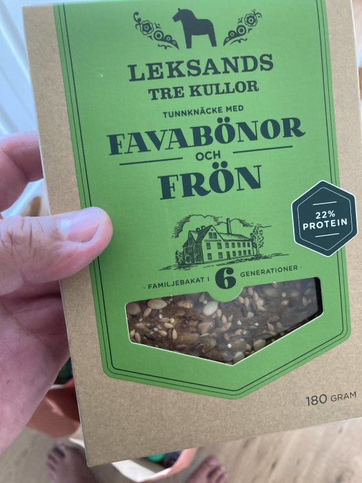 Hand håller ett paket Leksands knäckebröd med färgen favabönor och frön, närbild på förpackningen.