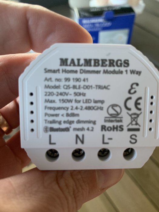 Hand håller upp en vit Malmbergs Smart Home Dimmer med tydliga anslutningsterminaler för elinstallation.