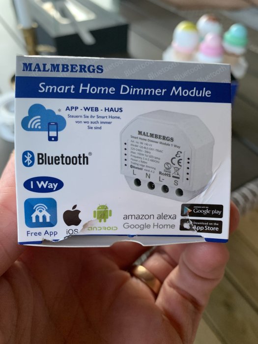 Hand håller en Malmbergs Smart Home Dimmer Module förpackning för styrning av lampor via app.