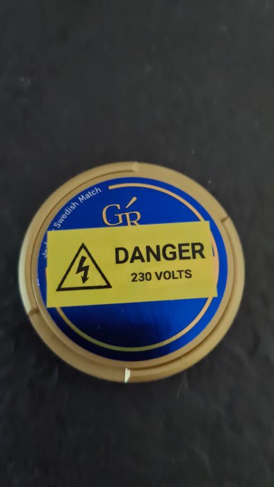 Rund varningsskylt med texten "DANGER 230 VOLTS" och en blixtsymbol, tejpad på en lockyta.