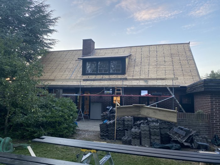 Hus med pågående takrenovering: hälften med takpapp och läkt, resten naket, plus material och pannor på marken.