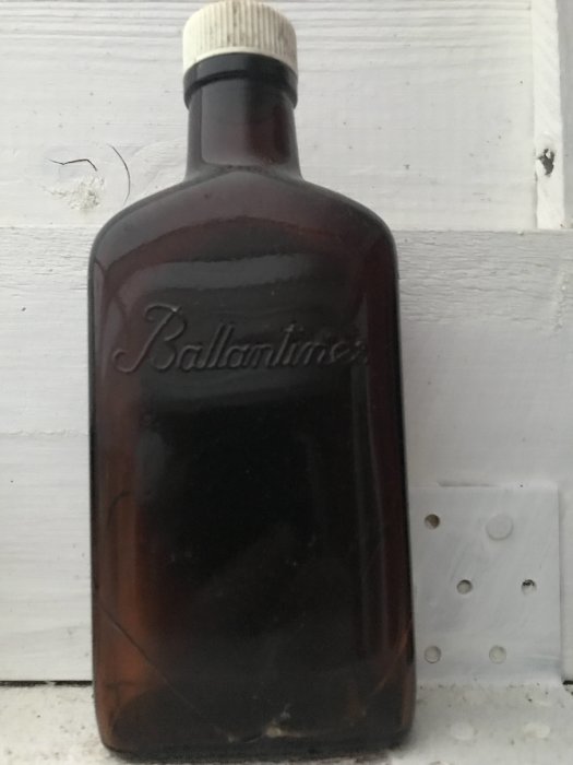 Gammal mörkbrun glasflaska med texten "Ballantine's" mot en vit vägg, hittad vid en stuga.