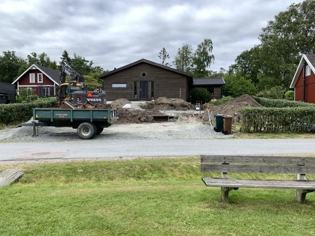 Grävmaskin på en uppgrävd tomt med grushögar framför ett brunt hus, erbjuder överskottsmaterial i Göteborg.