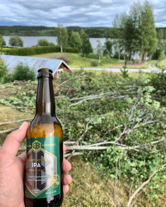 Hand håller en Jämtlands Bryggeri IPA-ölflaska framför en grönskande trädgård med sjöutsikt.