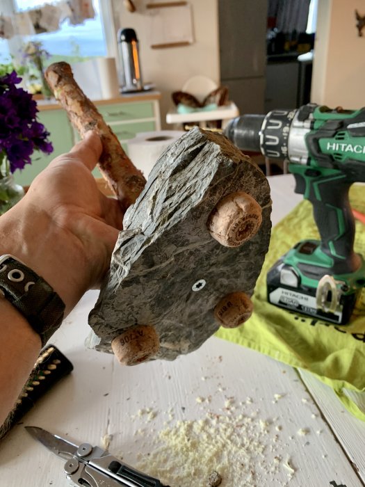 Hand håller en skiffersten med champagnekorkar som fötter bredvid en borrmaskin, för DIY pappersrullehållare.