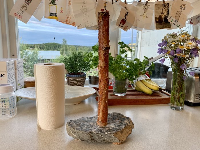 Hemmagjord pappersrullehållare av skiffer och björk på ett köksbord med utsikt över grönområde.