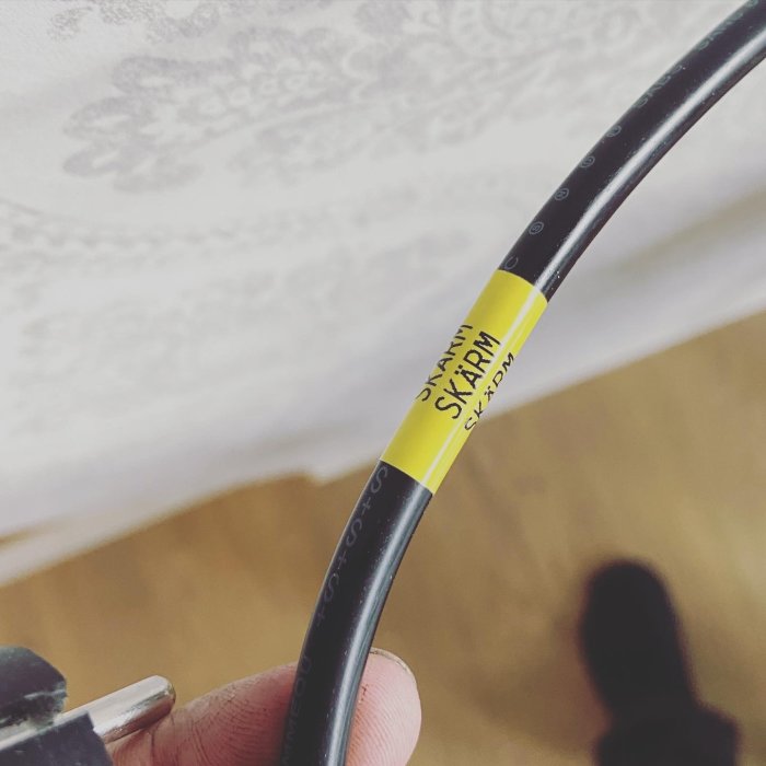 Hand håller en svart kabel med gul skarvkontakt märkt "SKARV SKÄRM".