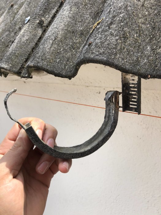 Hand håller en gammal rännkrok avsedd för 115 mm hängränna framför ett vågigt tak.