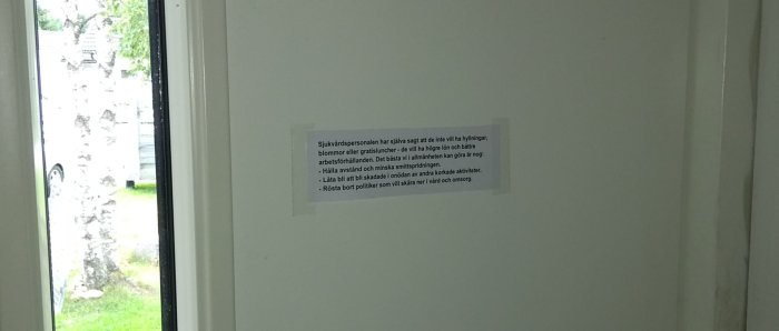 Lapp med text fäst på en vit dörr, synlig genom en glasruta, med suddig bakgrund av ett träd och en parkerad bil.