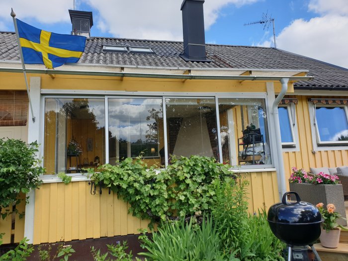 Uterum med stora fönster på gult hus, svensk flagga, vegetation och grill framför.