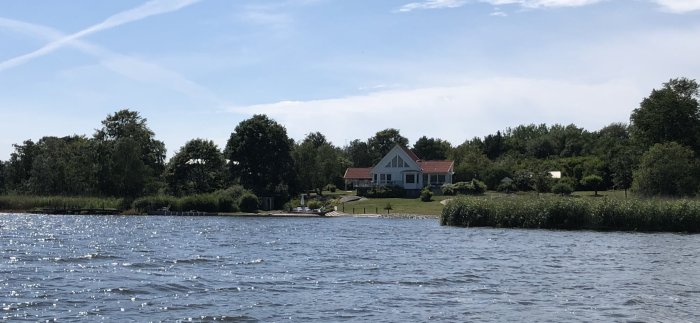 Vit hus med blåa fönsterluckor vid vattnet, omgiven av gröna träd och solglitter på vattenytan.