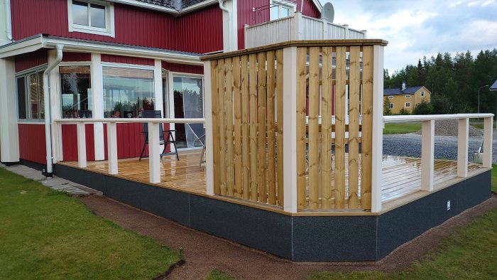 Rød og hvid bygning med terrasse af træ, omgivet af grønne områder, med et åbent træværksskur eller vindskærm.