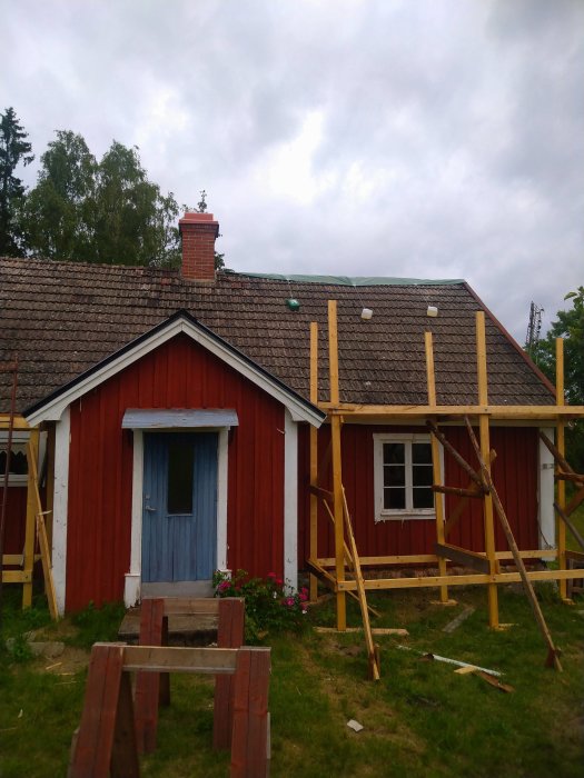 Rödmålat torp med gammalt spåntak under renovering, ställningar och reglar placerade för takriktning.