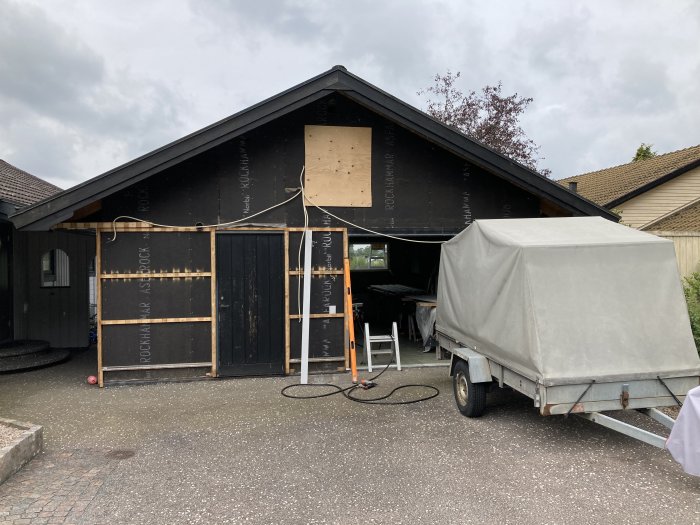 Renoveringsprojekt med delvis avmonterad ytterpanel och oskyddad väggstruktur på ett garage, med stege och släpvagn framför.