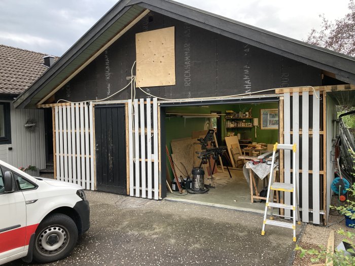 Garagebygge med pågående panelarbeten och verktyg synliga, delvis öppen garagedörr.