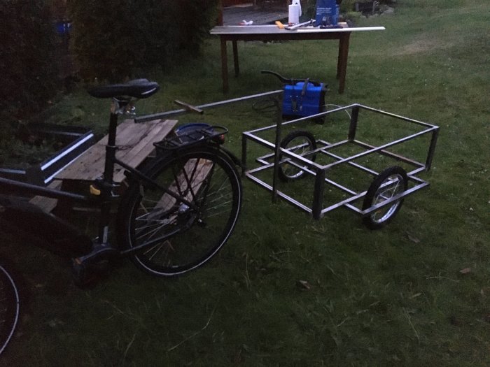 Svetsad metallram för cykelkärra med 16-tums hjul, bredvid en cykel och arbetsbord utomhus i skymningen.