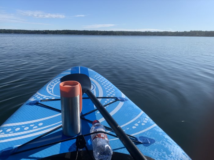 En stand-up paddleboard på lugnt vatten med en termos och vattenflaska ombord, skog i bakgrunden under klar himmel.