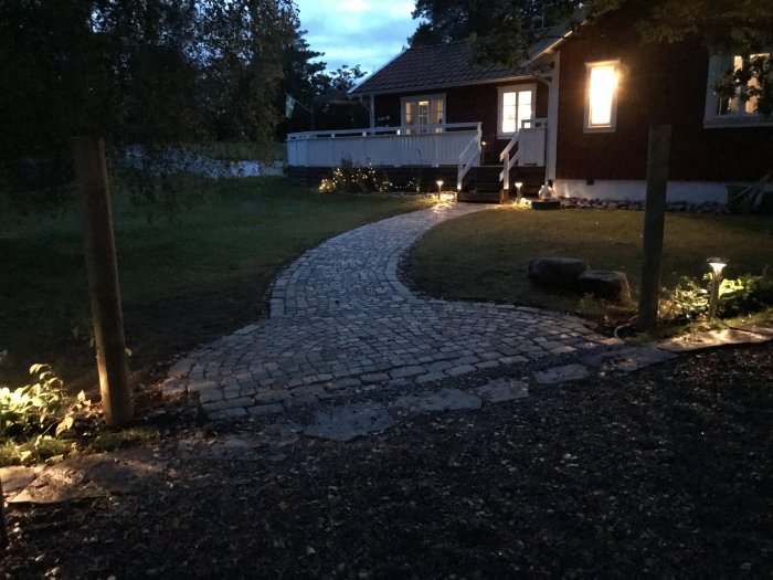 Gångväg med kullersten framför hus i skymning med belysning och trädgård.