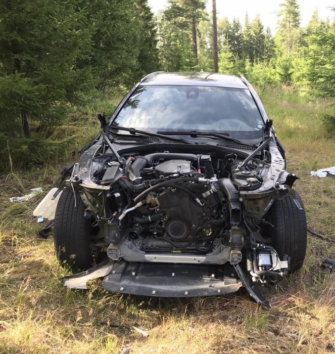 Nedmonterad främre del av en bil står i skogsmiljö, med motor och chassidelar exponerade.