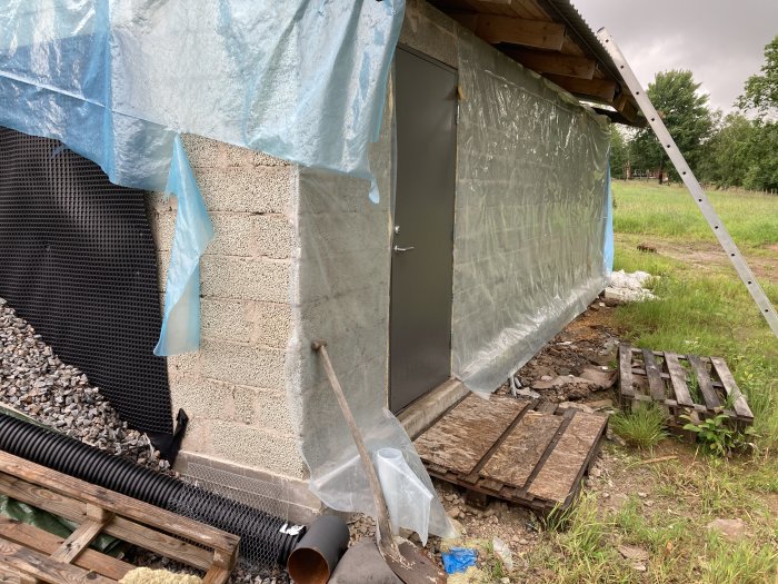 Oskyddat byggprojekt med öppen grund och presenningar under grå himmel.