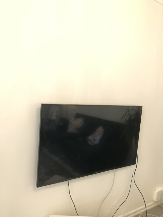 Flat-TV monterad på vit vägg med synliga kablar och reflektion av rummet i skärmen.