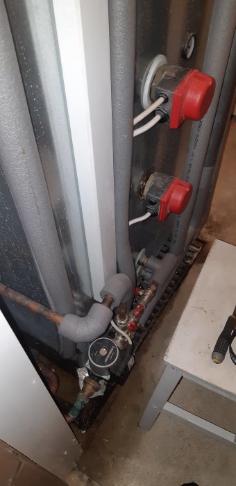 En bergvärmepump med isolerade rör och komponenter, inklusive en pump och ventil, i ett tekniskt utrymme.