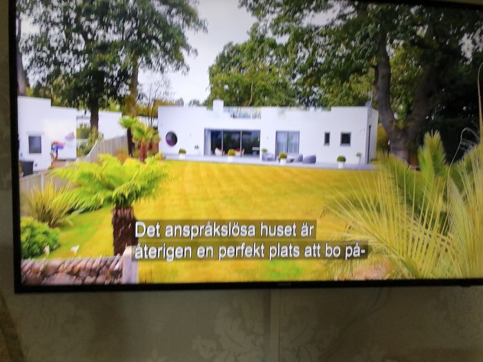 Vit, modern villa med platt tak och stora fönster, omgiven av en välskött trädgård och textremsa på TV-skärm.