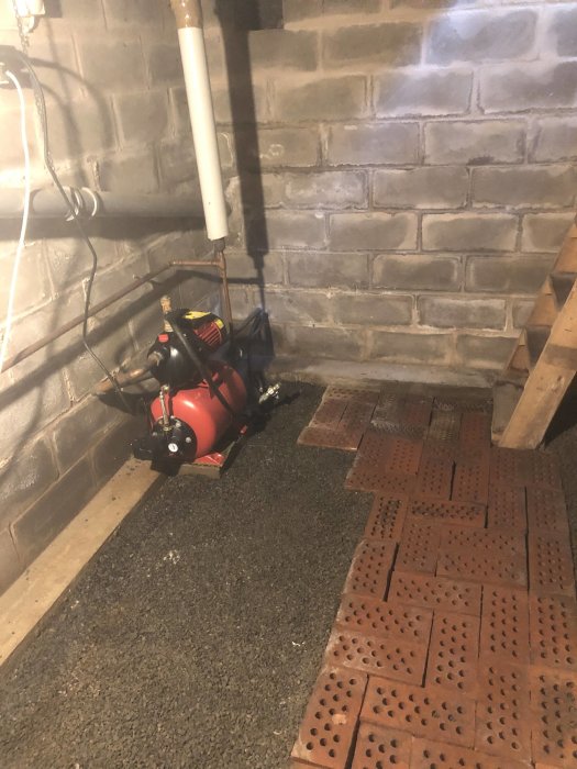 Källarrum med vattenpumpar på grusgolv och dräneringsplattor, ojämna väggar och synliga rör.