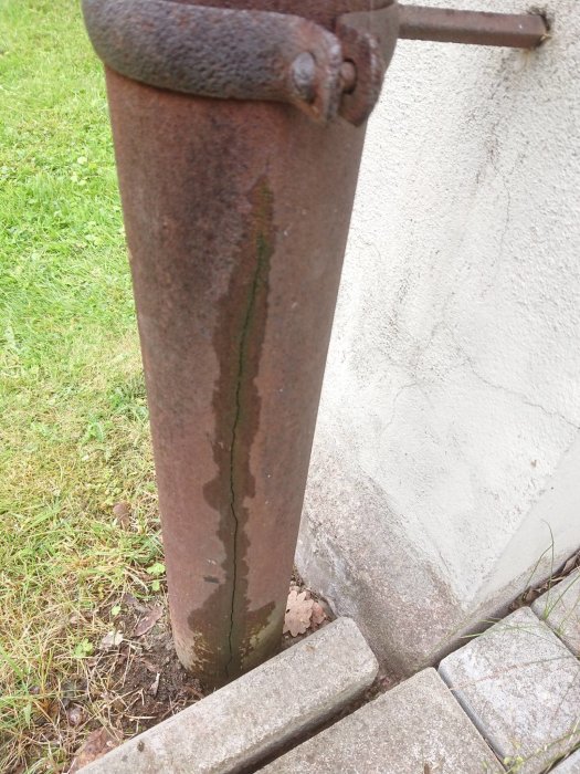 Järnrör med vertikal spricka bredvid husvägg och över betongrör, behöver ersättning.