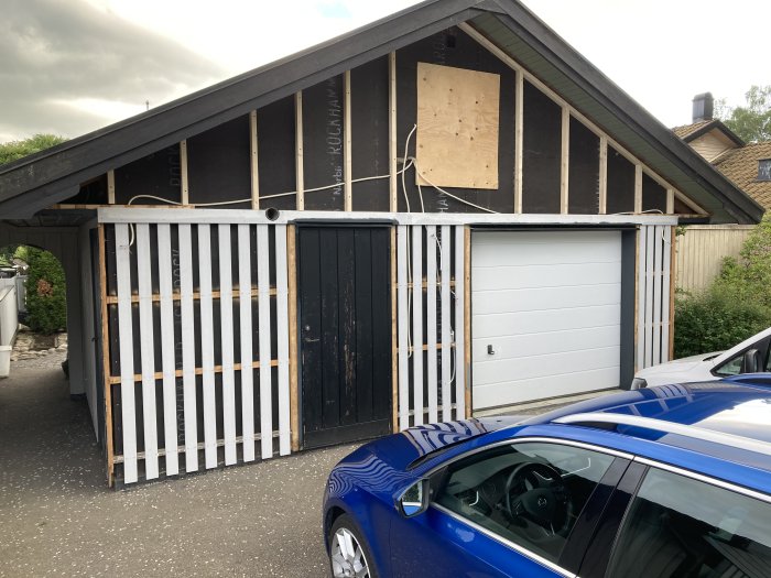 Ett garage under renovering med exponerad isolering och delvis monterade ytterpaneler bredvid en blå bil.