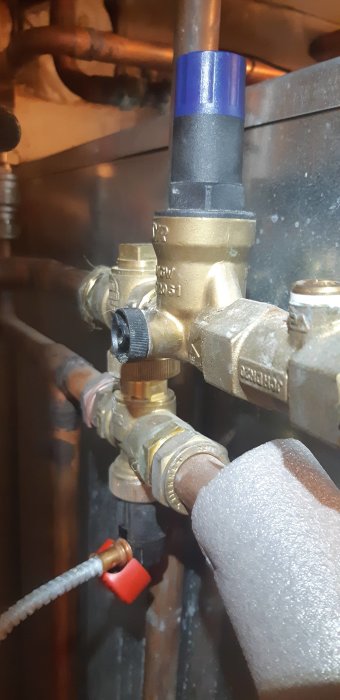 VVS-installation med kopparledningar och en blå säkerhetsventil, samt en röd avstängningskran och grå, isolerade rör.
