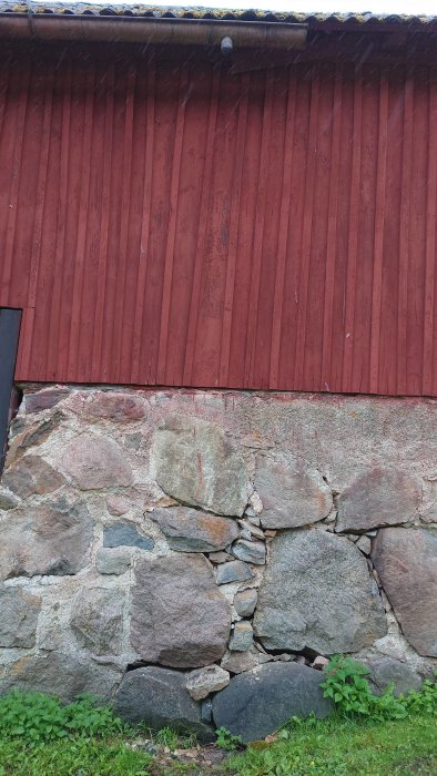 Grundmur med skadade delar och lösa stenar under en rödmålad trävägg och stupränna.