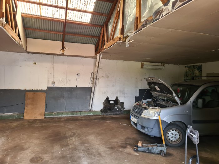 Ofärdigt garageinteriör med plåttak, en öppen bilhuva på en bil, och verkstadverktyg på betonggolv.
