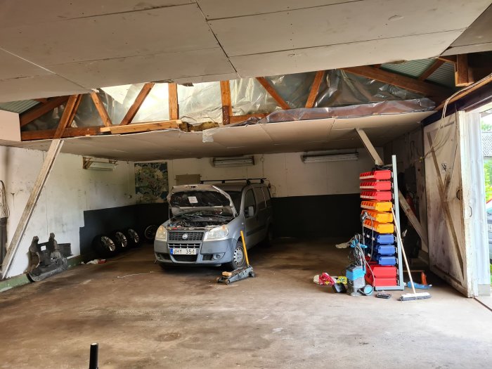 Interiör av verkstadsgarage med bil och verktyg, synliga takreglar och spånskivor på väggarna.