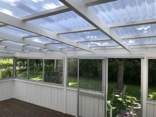 Renoverad terrass med nytt tak av transparent material, trägolv och vitmålade stolpar mot en trädgårdsbakgrund.