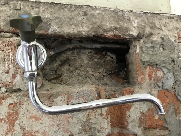 Kromad vattenkran framför en öppen murstock där röret syns gå in i väggen, möjlig koppling otydlig.
